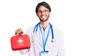 Handsome hispanic man wearing doctor coat holding first aid kit looking positive and happy standing and smiling with a confident smile showing teeth