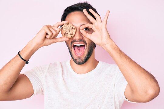 Handsome hispanic man holding cookie smiling happy doing ok sign with hand on eye looking through fingers