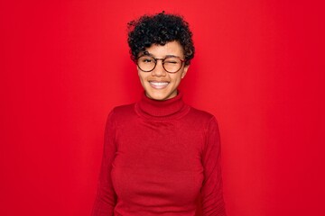 Young beautiful african american afro woman wearing turtleneck sweater and glasses winking looking...