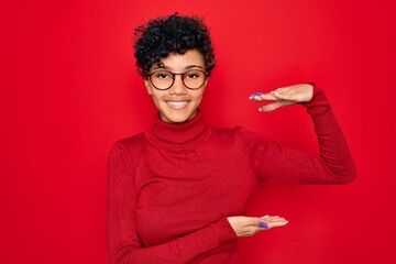 Young beautiful african american afro woman wearing turtleneck sweater and glasses gesturing with hands showing big and large size sign, measure symbol. Smiling looking at the camera. Measuring.