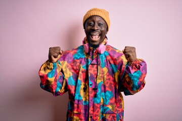 Obraz premium Young handsome african american man wearing colorful coat and cap over pink background celebrating surprised and amazed for success with arms raised and open eyes. Winner concept.