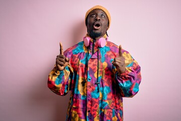 Young handsome african american man wearing colorful coat and cap over pink background amazed and surprised looking up and pointing with fingers and raised arms.