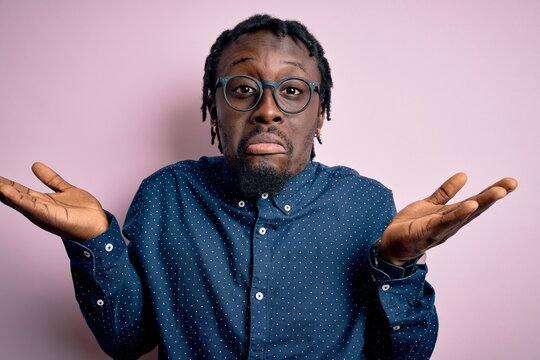 Young handsome african american man wearing casual shirt and glasses over pink background clueless and confused expression with arms and hands raised. Doubt concept.