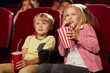 Portrait of two cute scared kids watching movie in cinema theater and eating popcorn, copy space