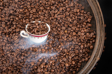 Roasted coffee on rustic sieve with smoke and a cup with black background,  top view