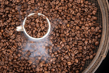 Roasted coffee on rustic sieve with smoke and a cup with black background,  top view