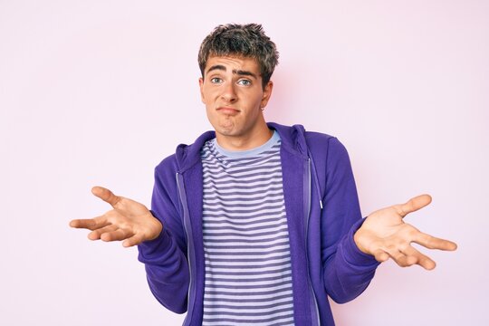 Young handsome man wearing casual purple sweatshirt clueless and confused expression with arms and hands raised. doubt concept.