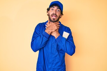Handsome young man with curly hair and bear wearing builder jumpsuit uniform shouting and suffocate because painful strangle. health problem. asphyxiate and suicide concept.