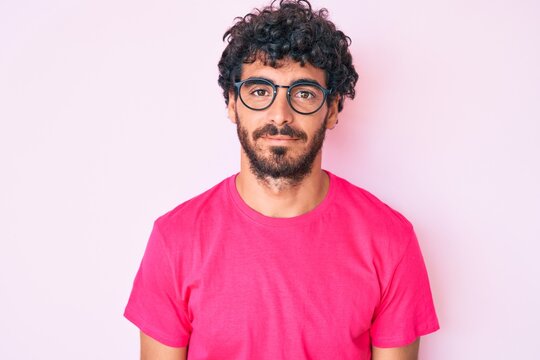 Handsome young man with curly hair and bear wearing casual clothes and glasses with serious expression on face. simple and natural looking at the camera.