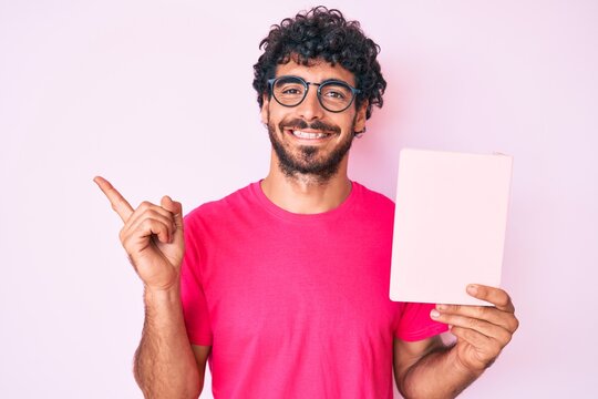 Handsome young man with curly hair and bear reading a  book smiling happy pointing with hand and finger to the side