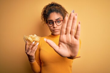 Young african american afro girl holding bowl with potatoes chips over yellow background with open hand doing stop sign with serious and confident expression, defense gesture