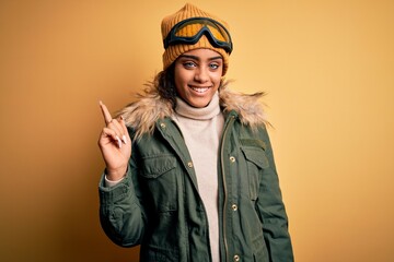 Young african american afro skier girl wearing snow sportswear and ski goggles with a big smile on face, pointing with hand finger to the side looking at the camera.