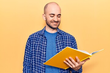 Young handsome bald man reading book looking positive and happy standing and smiling with a confident smile showing teeth