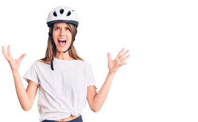 Young beautiful girl wearing bike helmet crazy and mad shouting and yelling with aggressive expression and arms raised. frustration concept.