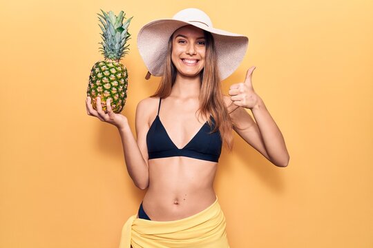 Young beautiful girl wearing bikini and hat holding pineapple smiling happy and positive, thumb up doing excellent and approval sign