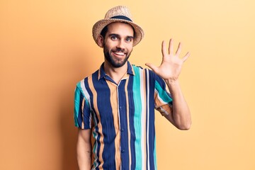 Young handsome man with beard wearing summer hat and shirt showing and pointing up with fingers number five while smiling confident and happy.