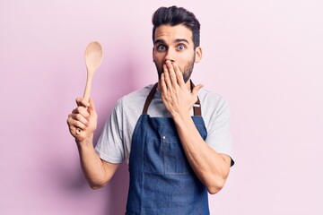 Young handsome man with beard wearing apron holding wooden spoon covering mouth with hand, shocked and afraid for mistake. surprised expression