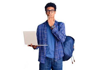 Young african american man holding student backpack using laptop covering mouth with hand, shocked and afraid for mistake. surprised expression