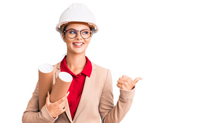 Young beautiful woman wearing architect hardhat and glasses holding blueprints pointing thumb up to the side smiling happy with open mouth