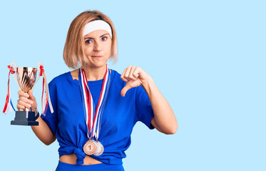 Young blonde woman holding champion trophy wearing medals with angry face, negative sign showing...