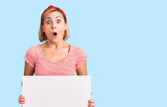 Young blonde woman holding blank empty banner scared and amazed with open mouth for surprise, disbelief face