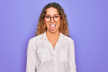 Young beautiful woman with blue eyes wearing casual shirt and glasses over purple background sticking tongue out happy with funny expression. Emotion concept.