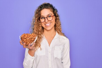 Obraz premium Young beautiful blonde woman with blue eyes holding bowl with healthy almond snack with a happy face standing and smiling with a confident smile showing teeth