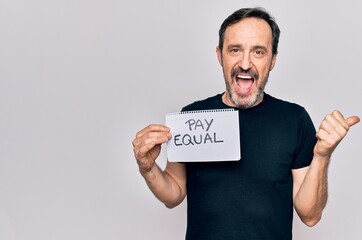 Middle age handsome man asking for social rights holding paper with pay equal message pointing thumb up to the side smiling happy with open mouth