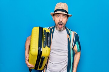 Middle age handsome tourist man on vacation holding cabin bag over isolated blue background scared and amazed with open mouth for surprise, disbelief face
