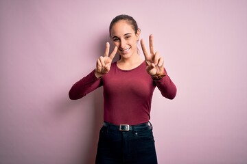 Obraz na płótnie Canvas Young beautiful woman wearing casual t-shirt standing over isolated pink background smiling looking to the camera showing fingers doing victory sign. Number two.