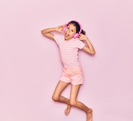Adorable hispanic child girl smiling happy. Jumping with smile on face listening to music using pink headphones over isolated background