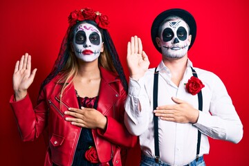 Couple wearing day of the dead costume over red swearing with hand on chest and open palm, making a loyalty promise oath
