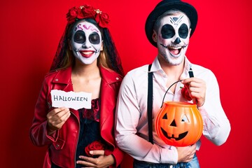 Couple wearing day of the dead costume holding pumpking and halloween paper smiling and laughing hard out loud because funny crazy joke.