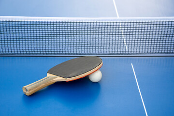 Black wooden racket for ping pong and white ball lying on the blue table next to the net