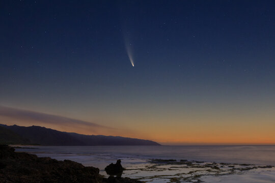 Comet Neowise at dawn over coastal tide pools in the foreground and wisps of clouds over the mountains.
