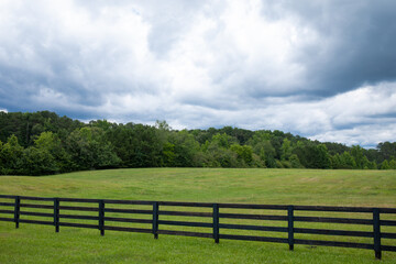 Storm clouds over a brilliant green field, black rail fence in front with treeline on the horizon, horizontal aspect