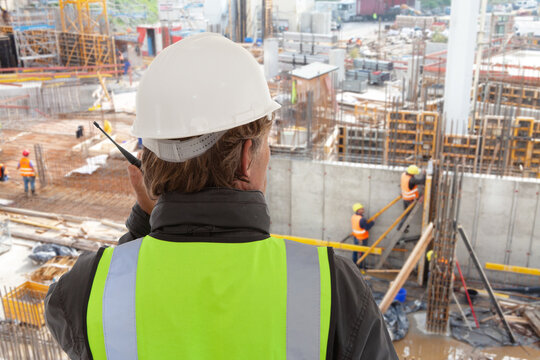 construction worker or civil engineer or architect or foreman with hard hat and safety vest at major construction site formworks supervising with walkie talkie