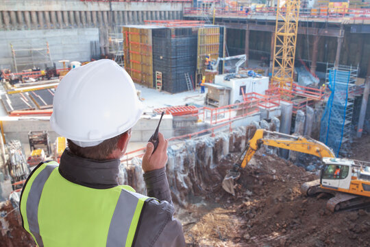 construction worker or civil engineer or architect or foreman with hard hat and safety vest at major construction site with crane and digger supervising with walkie talkie