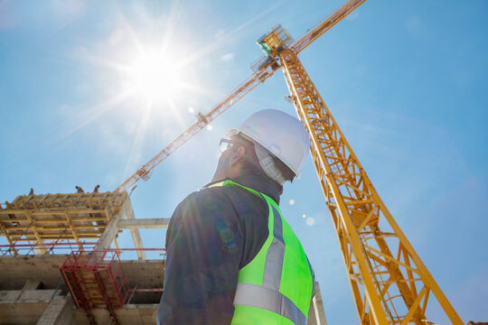 architect or construction worker or civil engineer or foreman with hard hat and safety vest on construction site  with crane and sun flare