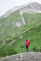 Naklejka premium Vertical shot back view of female hiker with backpack, enjoying the view of beautiful hills covered with grass. Woman with trekking sticks looking at majestic mountain landscape. Concept of hiking.
