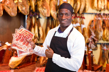 Happy African American seller working in meat shop, recommending delicious iberian ham..