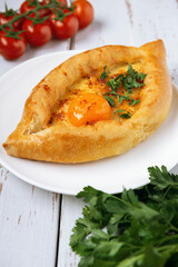 Adjarian Khachapuri – Georgian cheese bread decorated with tomatoes and parsley on white plate and Provence style background. Baked open pie with cheese and yolk filling.