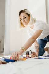 Beautiful young blonde woman getting ready to paint at home. Choosing instrument to paint. many painting oil colors and paint brushes on the floor.