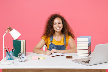 Smiling young african american girl employee in office sit work at desk isolated on pink background. Achievement business career. Education in school university college concept. Write in notebook.