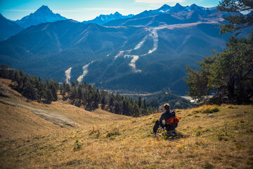 Fototapeta premium A tourist sits in a clearing and looks into the distance. Nearby are large conifers, below the trees there is dry grass and fallen cones, mountains are visible in the distance.