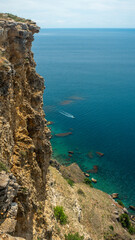 A sheer cliff overlooking the Black Sea, a kayak rides in the distance, a calm turquoise blue sea below the edge. Light bright weather, blue sky without clouds.