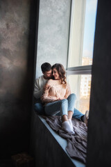 Happy couple in love in winter sweaters and jeans, sit on a wide windowsill by the window, hug. A young man kisses a beautiful curly brown-haired girl on the cheek. Date by the fireplace in  cozy home