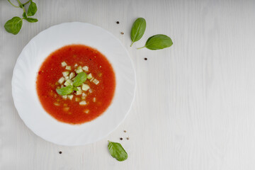 White plate with red gazpacho soup made of pureed tomatoes and other vegetables standing on light wooden table at kitchen surrounded by basil leaves. Image with copy space, horizontal orientation
