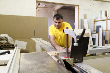 Carpenter cutting a piece of wood for furniture in his woodwork workshop, using a table saw, and wearing yellow tshirt.