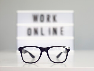 Glasses on desk and blurred sign with words WORK ONLINE against grey background. Home based work concept